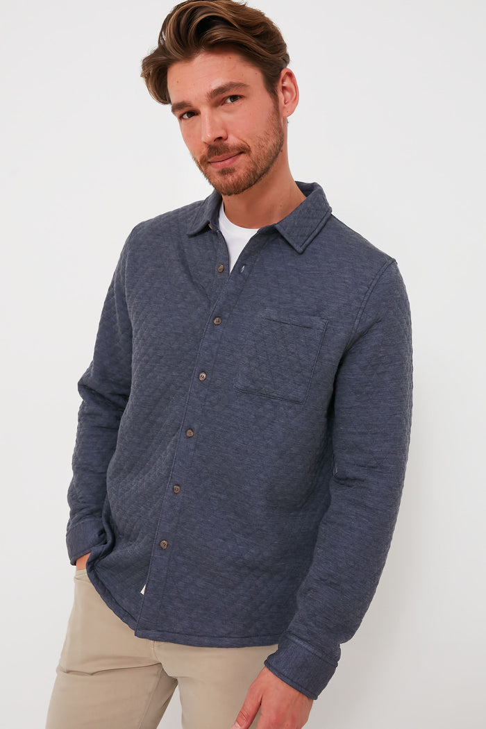 A man with light brown hair and a beard models a blue Marine Layer quilted button-up shacket and beige pants, highlighting an eco-friendly look.