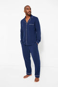 A man stands barefoot, wearing a navy pajama set with white piping made from luxurious Peruvian Pima cotton by Petite Plume.