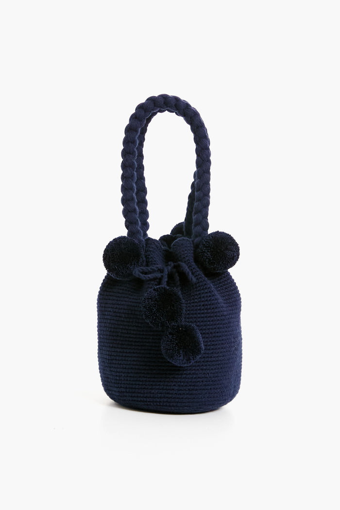 The Marina Orth Foundation navy blue knit bucket bag features a braided handle and playful pom-pom accents, blending artisan elegance and style—an ideal woven handbag for any occasion.