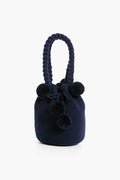 The Marina Orth Foundation navy blue knit bucket bag features a braided handle and playful pom-pom accents, blending artisan elegance and style—an ideal woven handbag for any occasion.