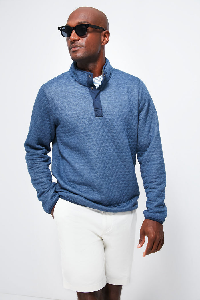 A man in Marine Layer black sunglasses, a diamond-quilted sweatshirt, and white shorts stands with one hand in his pocket.