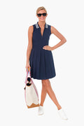 A woman in a TORY SPORT navy performance pleated golf dress with a striped collar, white sneakers, sunglasses, and carrying a large cream tote bag with rainbow straps.