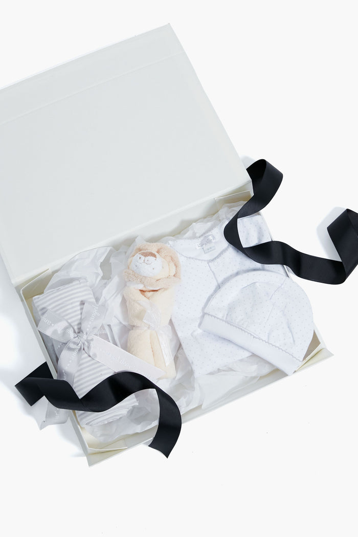 The Tuckernuck Tots baby gift bundle with a black ribbon includes a striped box, plush toy, white baby hat, and white polka-dot outfit on tissue paper—ideal as a newborn gift set or for baby showers.