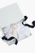 The Tuckernuck Tots baby gift bundle with a black ribbon includes a striped box, plush toy, white baby hat, and white polka-dot outfit on tissue paper—ideal as a newborn gift set or for baby showers.