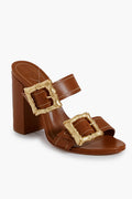 Schutz's brown Enola Sandals feature block heels, two wide straps with large gold buckles, and white stitching—stylish statement shoes for any occasion.