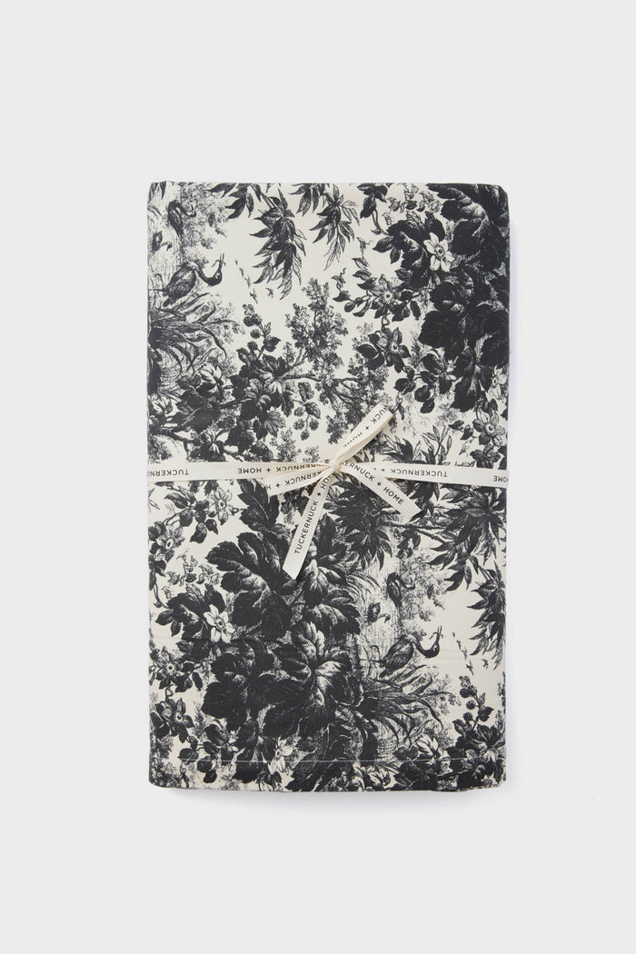 A folded black and white floral cotton Tuckernuck Home tablecloth, tied with a cream-colored ribbon, ready to use on your rectangle table.
