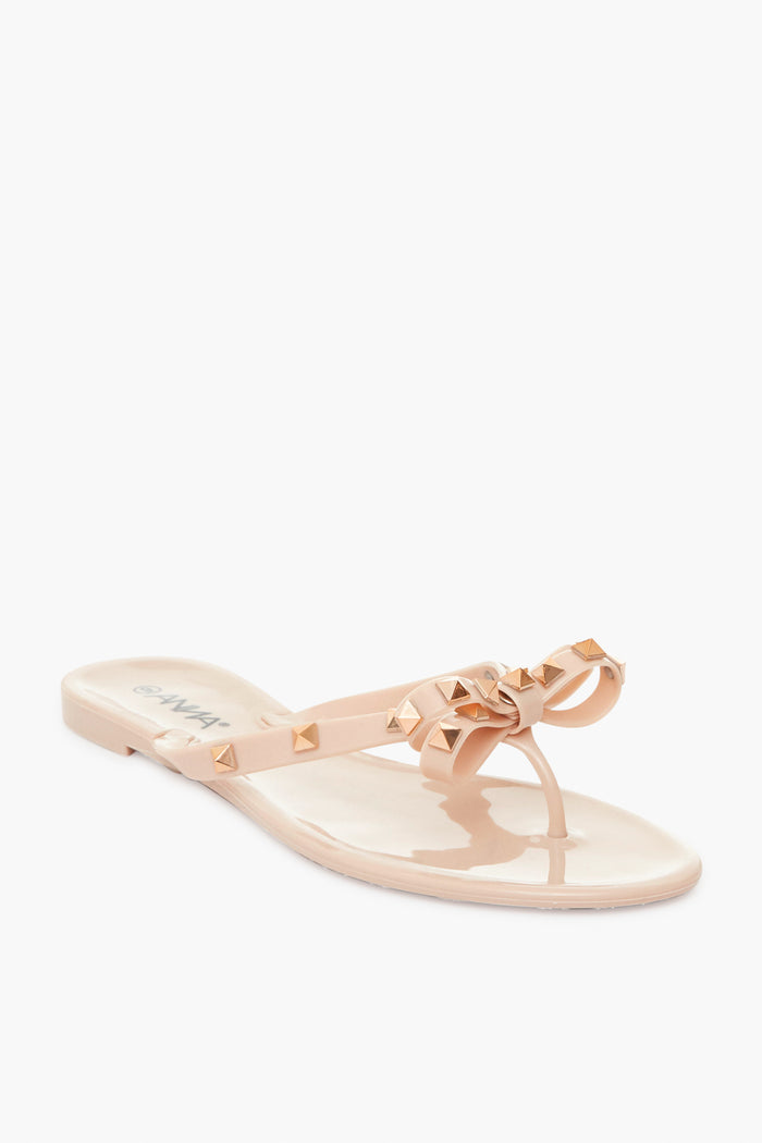 Anina Beige Nude Jelly Bowtie Sandals feature a glossy PVC finish, gold-tone pyramid studs, bow detail, and a single toe post.