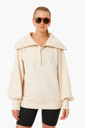 A person in Varley sunglasses, a beige half zip pullover with a wide collar, and black cycling shorts stands facing forward, blending performance fashion with casual style.