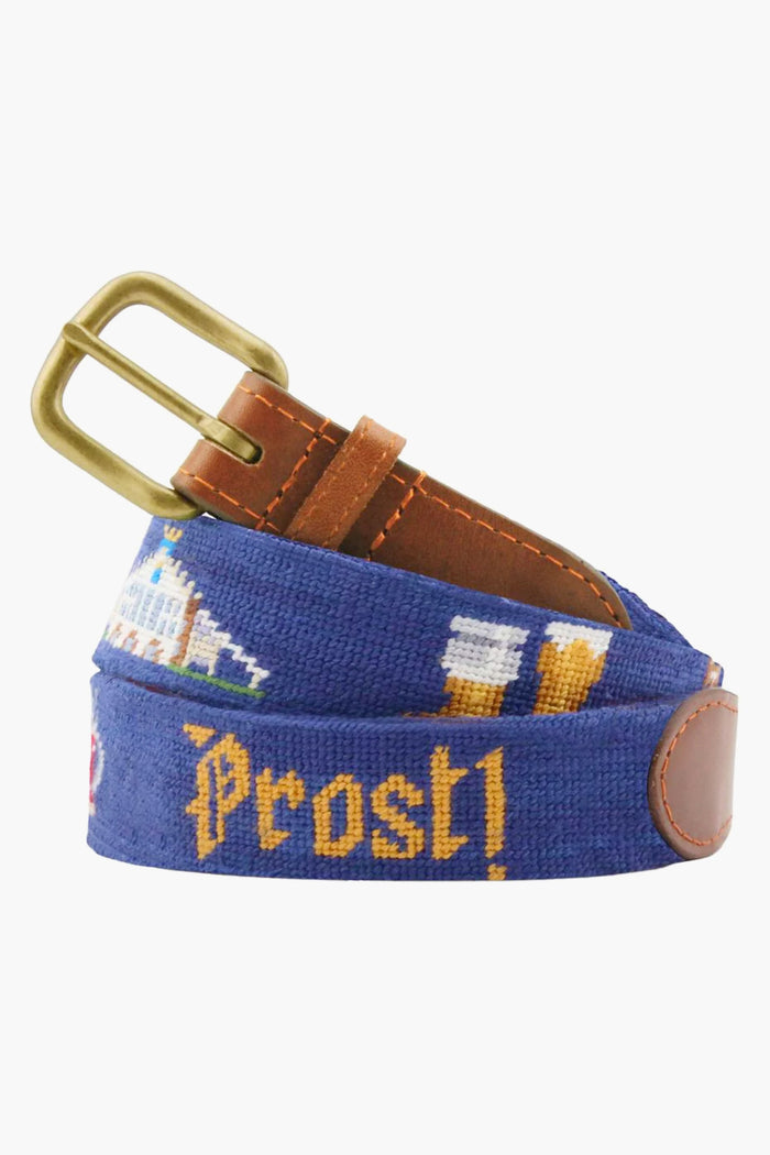 A Smathers and Branson blue needlepoint belt with brass buckle, leather accents, and hand-stitched pixel art of beer glasses, a building, and yellow "Prost!"—ideal for Oktoberfest celebrations.