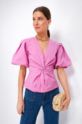A woman in a SIMKHAI pink cotton poplin puff sleeve top and blue jeans holds a woven clutch, gazing slightly to the side.