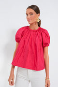 A woman wears the Ulla Johnson bright pink Orchid Lora Top with puff sleeves and white pants, accessorized with gold flower earrings for a romantic blouse look.