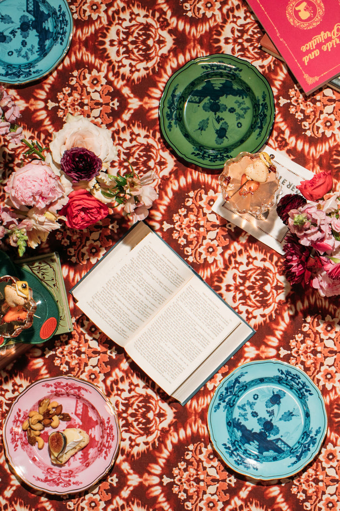 A patterned tablecloth displays colorful Ginori 1735 porcelain salad plates with snacks, an open book, a closed red book, and floral arrangements.