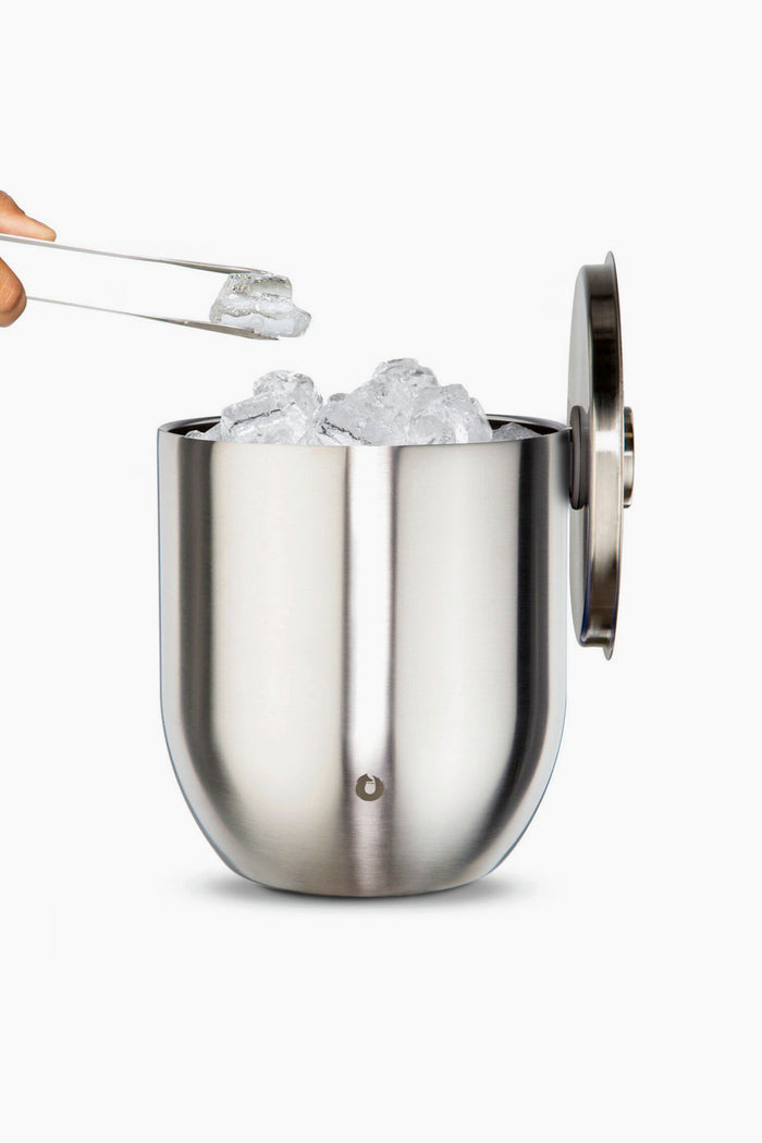 A Snowfox double-walled stainless steel ice bucket filled with ice cubes sits open, as tongs lift an ice cube above the insulated barware container.