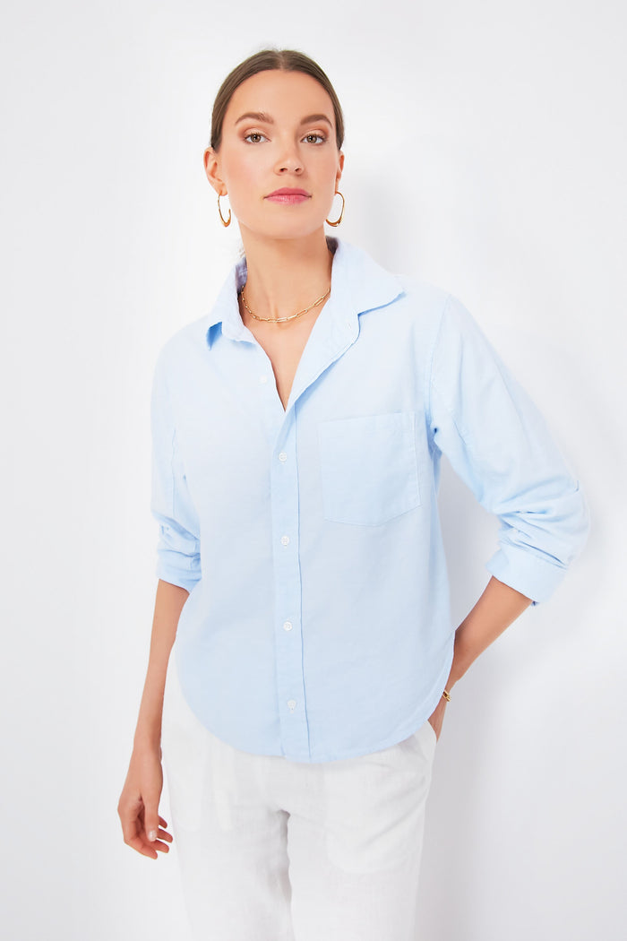A woman wears a Citizens of Humanity Oxford Blue Kayla Shrunken Shirt with rolled sleeves, paired with white pants, gold hoop earrings, and a necklace—ideal for those who value sustainable fashion.