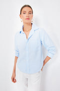 A woman wears a Citizens of Humanity Oxford Blue Kayla Shrunken Shirt with rolled sleeves, paired with white pants, gold hoop earrings, and a necklace—ideal for those who value sustainable fashion.