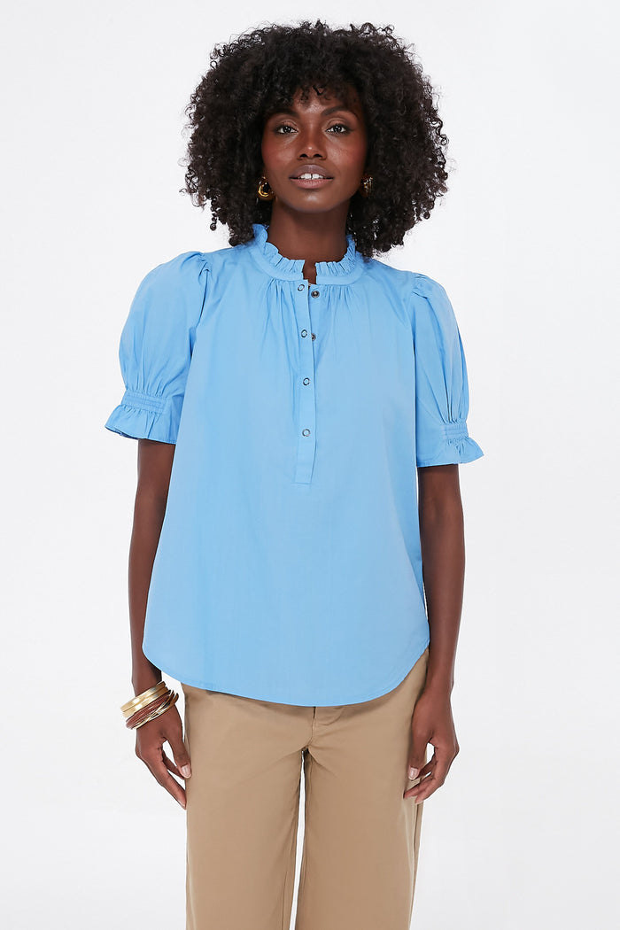 A woman wears an Apiece Apart light blue ruffle collar blouse made from organic cotton, paired with beige pants.