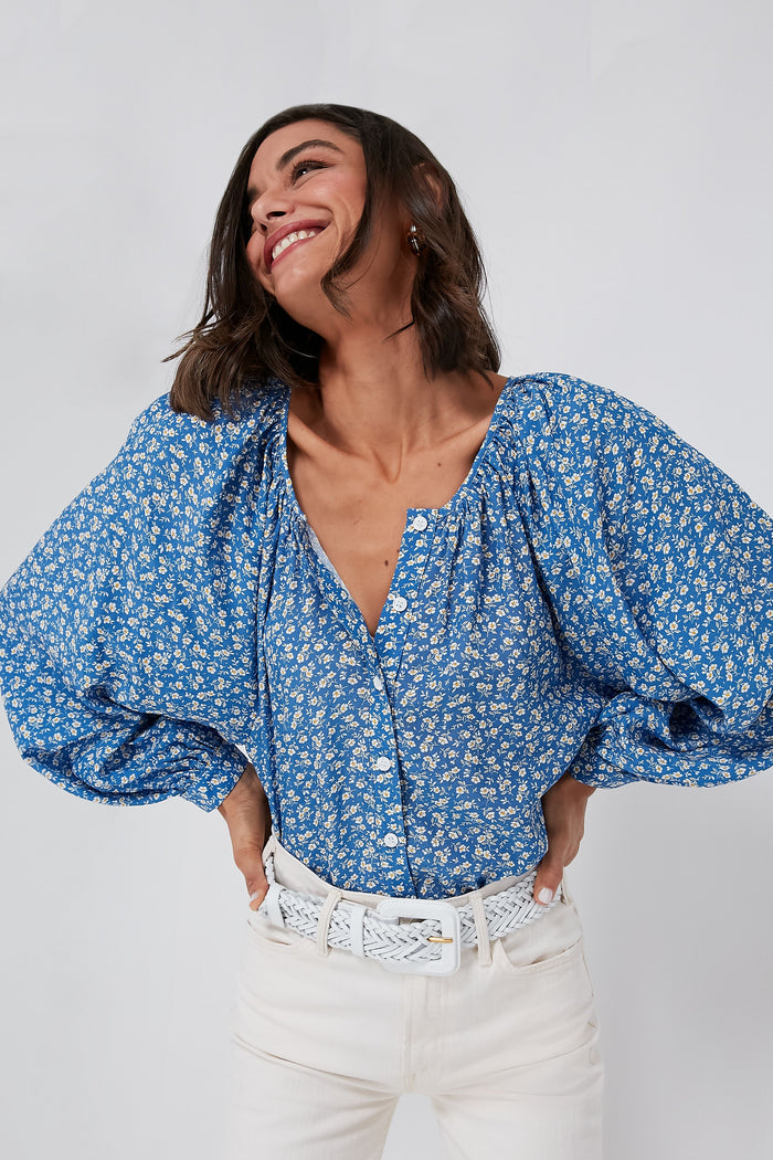A woman smiles with her hands on her hips, wearing Emerson Fry’s blue boho floral blouse with puffed sleeves and white pants.