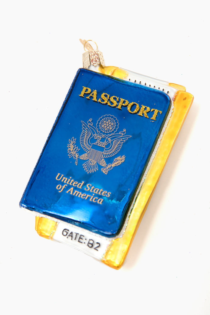 A hand-crafted Old World Christmas ornament, shaped like a blue U.S. passport, sits atop a yellow boarding pass labeled "GATE: B2"—a perfect collectible for holiday décor enthusiasts.