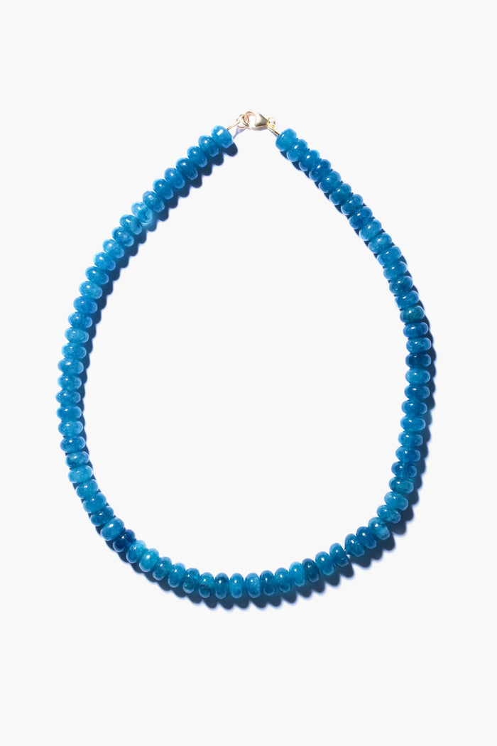 This handmade angelite necklace by Theodosia features round, bright blue beads with a small gold clasp, elegantly arranged in an oval shape—a stunning piece of jewelry.