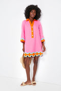 A woman wears the Tuckernuck x Atlantic-Pacific Collier Dress, a pink linen mini with orange, white, and blue geometric trim. She holds a straw hat and wears light-colored sandals.