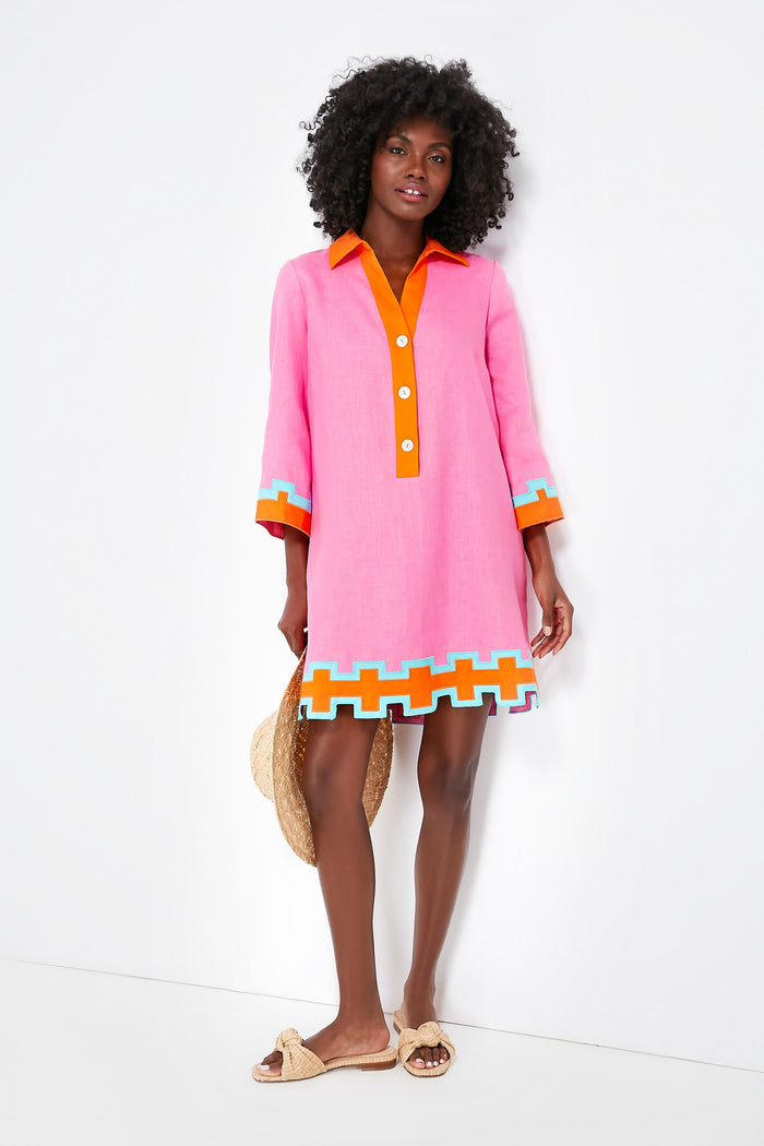 A woman wears the Tuckernuck x Atlantic-Pacific Collier Dress, a pink linen mini with orange, white, and blue geometric trim. She holds a straw hat and wears light-colored sandals.