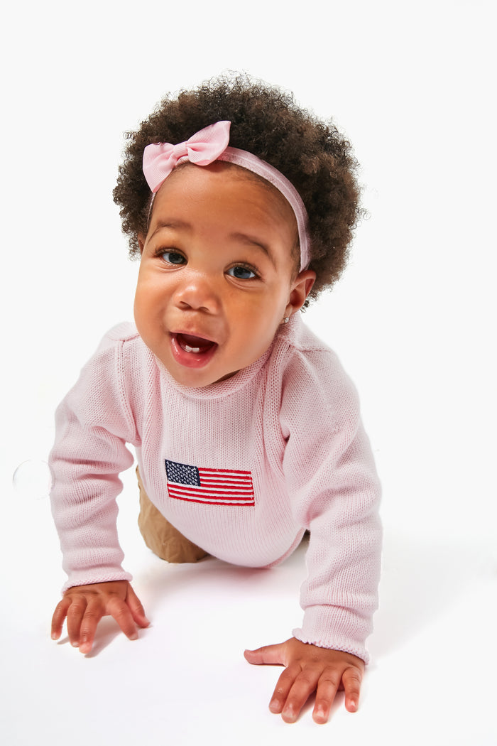 A baby in a pink, machine washable cotton sweater by A Soft Idea, featuring an American flag, and a pink headband crawls forward.