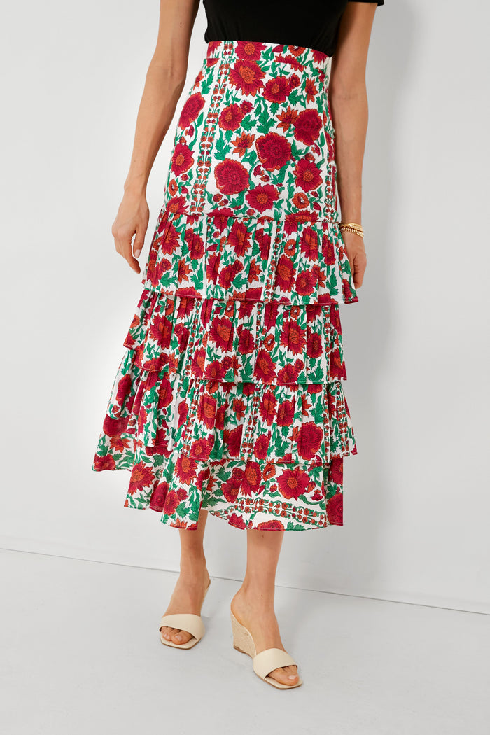 A woman wears a Ro's Garden black top and a tiered floral-print maxi skirt with red and green patterns, pleated ruffles, and light sandals—an ideal choice for luxury travel wear.