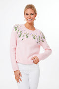 A woman wears the Hyacinth House Pink Floral Embroidered Sweater, featuring delicate embroidery on the chest and shoulders, paired with white pants as she stands smiling in a stylish, modern setting.