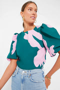 A woman wears the Hyacinth House Penelope Top—a green puff sleeve blouse with pink floral print—paired with light blue high-waisted jeans and gold jewelry, adding a fresh, stylish touch as she looks to the side.
