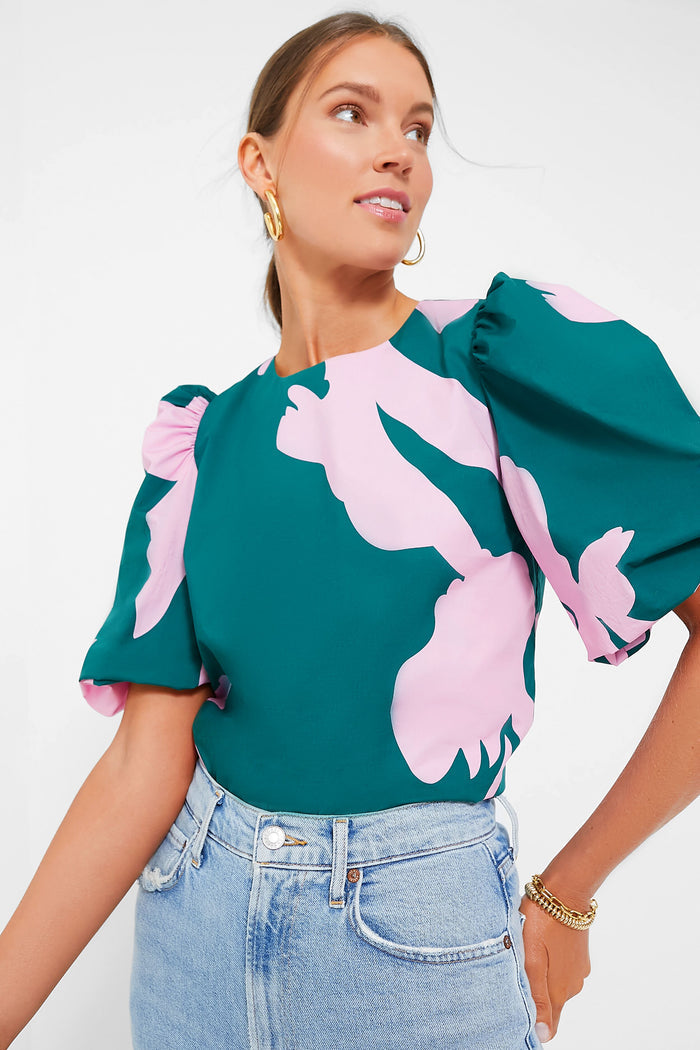 A woman wears the Hyacinth House Penelope Top—a green puff sleeve blouse with pink floral print—paired with light blue high-waisted jeans and gold jewelry, adding a fresh, stylish touch as she looks to the side.