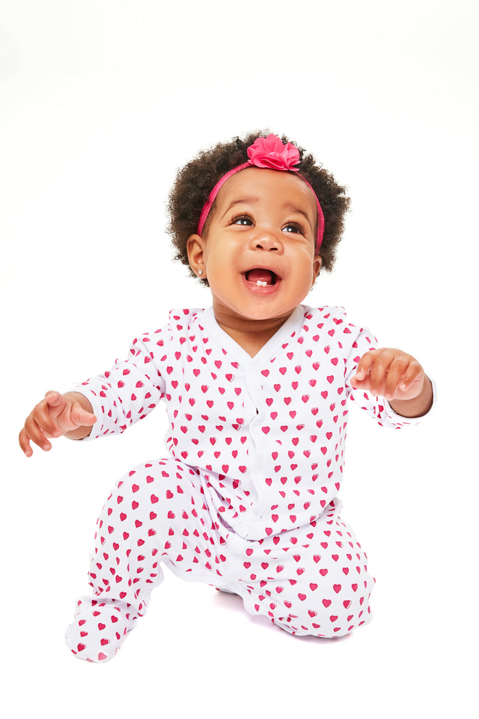A baby in Roller Rabbit heart-print pima cotton pajamas with a pink headband sits, smiling and looking upward.