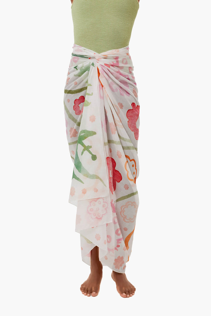 A person in a green sleeveless top and a white ASHA brand pareo adorned with colorful floral motifs, standing barefoot.