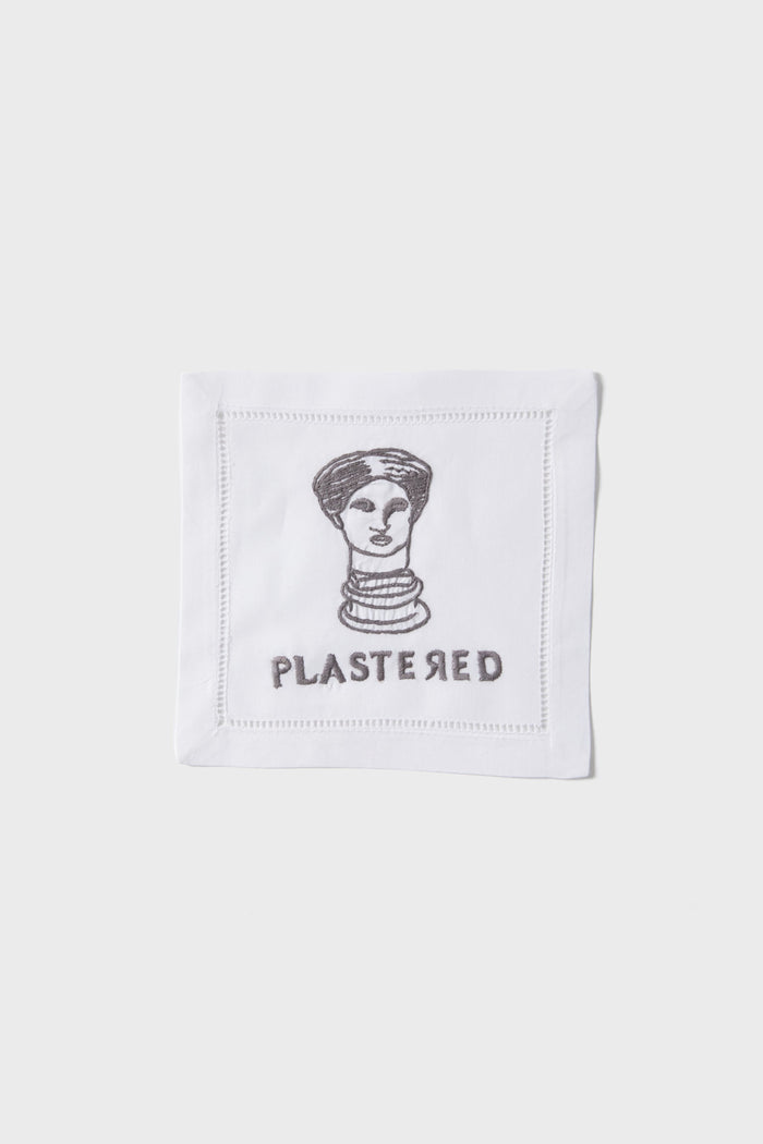 The August Morgan white linen cocktail napkin features a cheeky black line drawing of a woman’s head above “PLASTERED”—a fun hostess gift or playful addition to your next gathering.