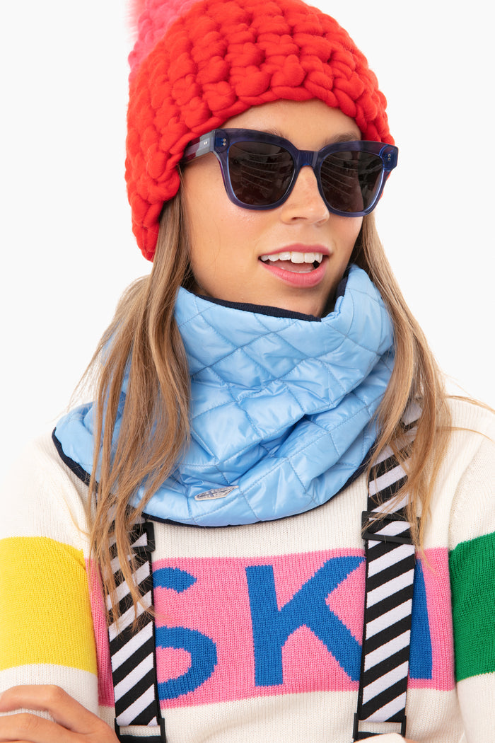 A person wears a red knit hat, dark sunglasses, a blue quilted scarf, and a Powder Puff Collection cashmere pullover sweater with SKI in colorful letters—a luxe winter accessory crafted from sustainable materials.
