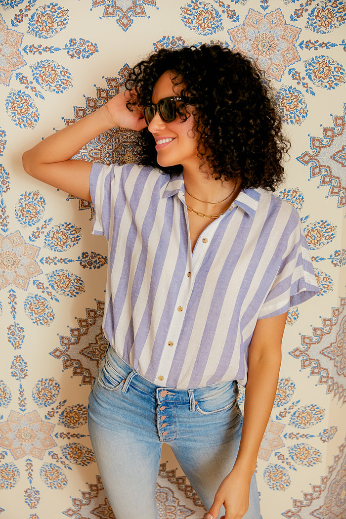A woman wearing sunglasses, a blue and white striped short-sleeve shirt, and light blue jeans stands smiling, showcasing a stackable jewelry look with Electric Picks Python Necklace.