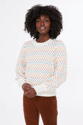 A person wears a Marine Layer eco-friendly sweater featuring a rainbow dot motif and brown pants, hands clasped in front.