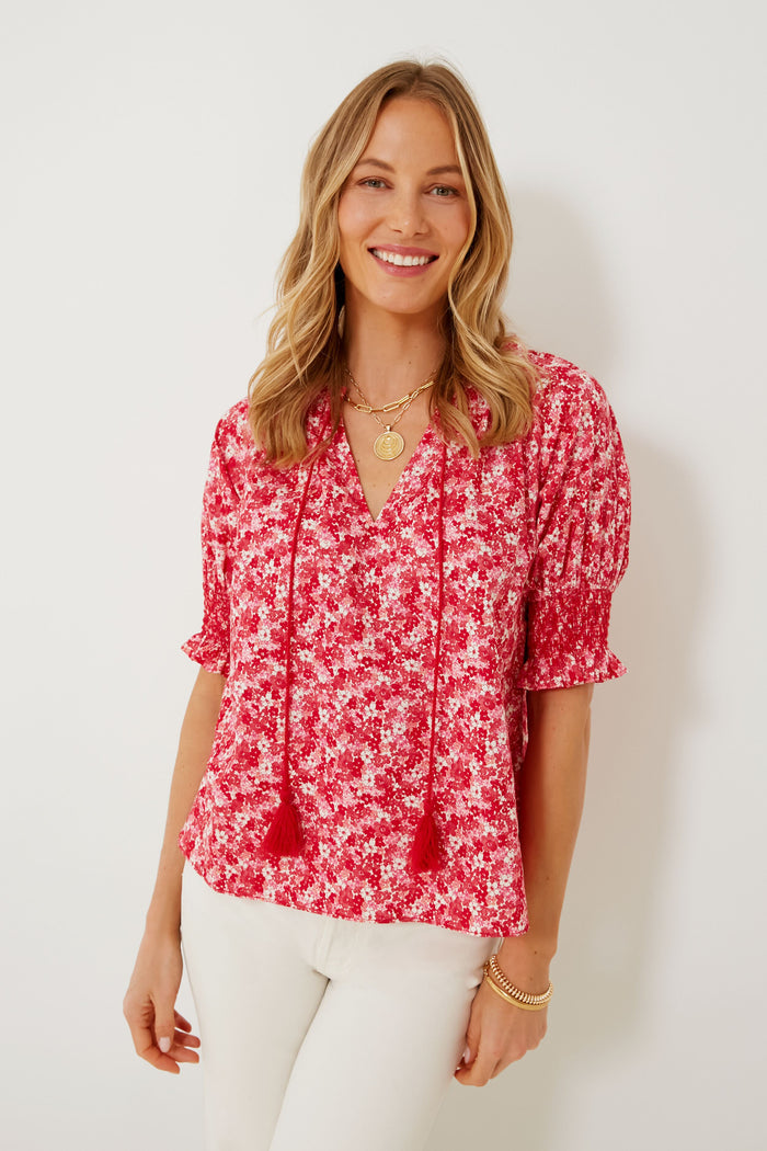 A woman smiles at the camera, wearing a red and white floral Hyacinth House Colleen Blouse with tassels and white pants.
