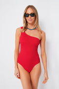 A woman in a Hunza G red asymmetrical one-piece swimsuit, black sunglasses, and a beaded necklace stands confidently, showcasing the brand’s signature crinkle-stretch fabric and chic design details.