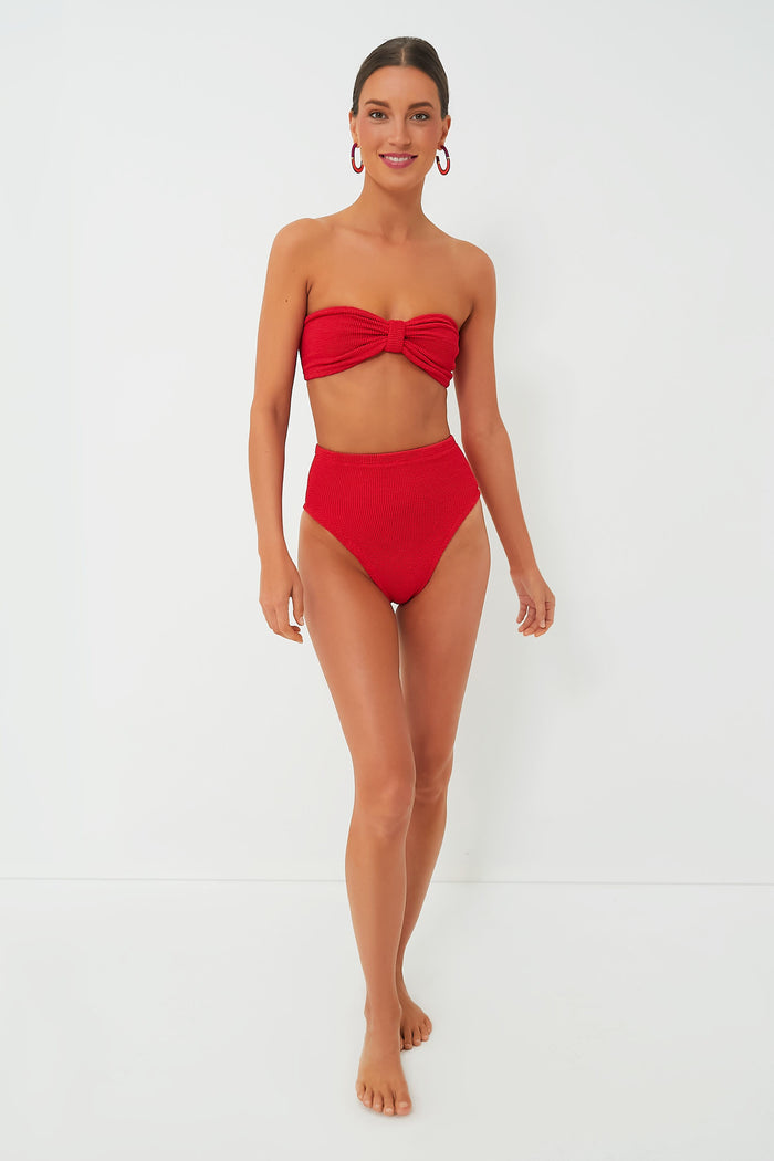 A woman stands barefoot in a Hunza G red crinkle-stretch bikini featuring a bow detail and retro high-waisted bottoms, paired with red hoop earrings.