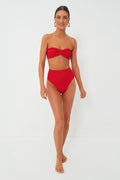 A woman stands barefoot in a Hunza G red crinkle-stretch bikini featuring a bow detail and retro high-waisted bottoms, paired with red hoop earrings.