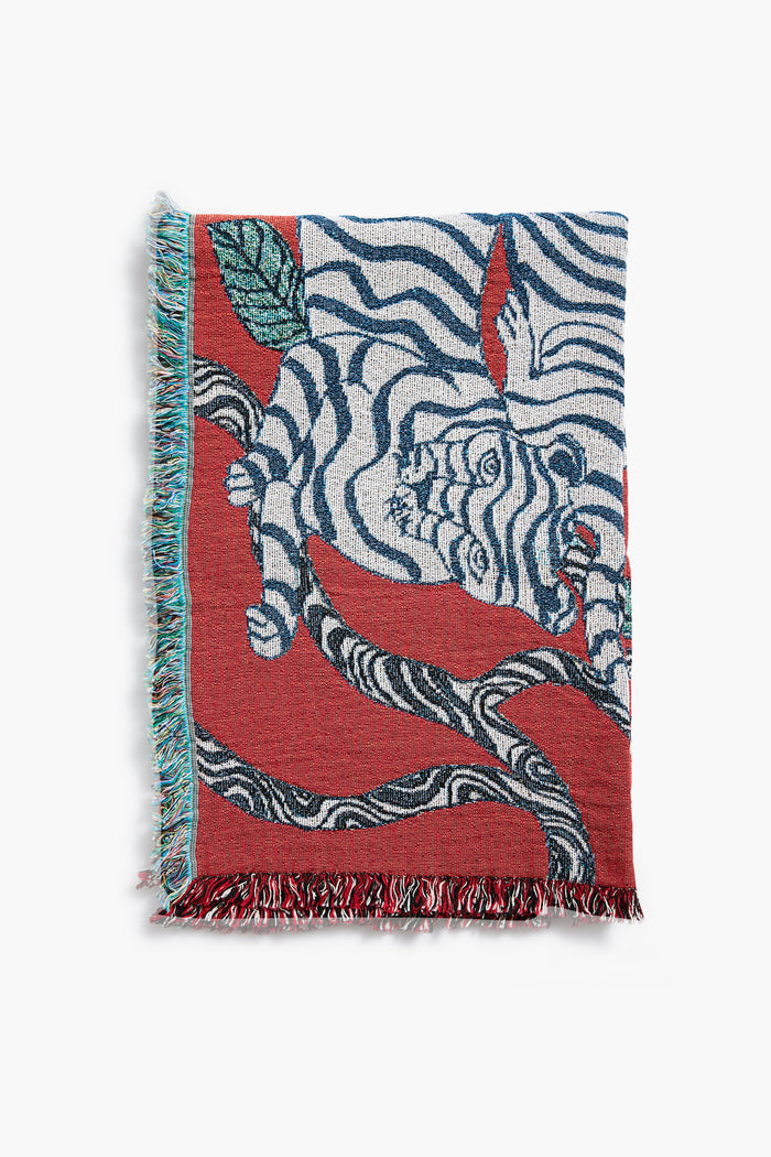 The Olivia Wendel Red Tigers Blanket features a white tiger with black stripes on a tree branch among green leaves, set against red hues. Made from 100% US-grown cotton, it’s finished with multicolored fringe edges.