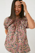 A woman smiles and touches her hair while wearing an Oliphant boho-chic pink floral short-sleeve organic cotton blouse, paired with a gold wristwatch.