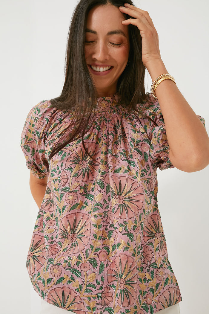 A woman smiles and touches her hair while wearing an Oliphant boho-chic pink floral short-sleeve organic cotton blouse, paired with a gold wristwatch.