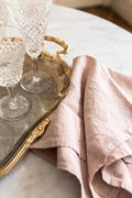 Three crystal wine glasses rest on an ornate silver tray beside a folded Madame de la Maison Rose Linen Napkin atop marble, adding a touch of sustainable elegance to your table setting.