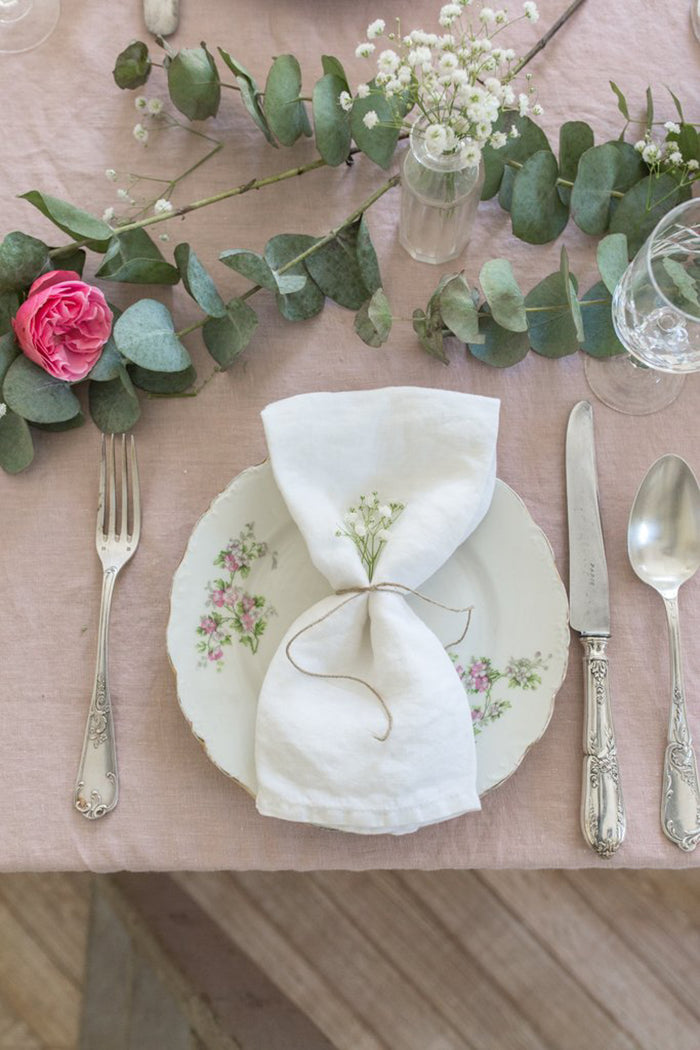 A table setting featuring a vintage floral plate, folded white napkin tied with string, silverware, and a pink rose with greenery on a Madame de la Maison flax linen tablecloth.