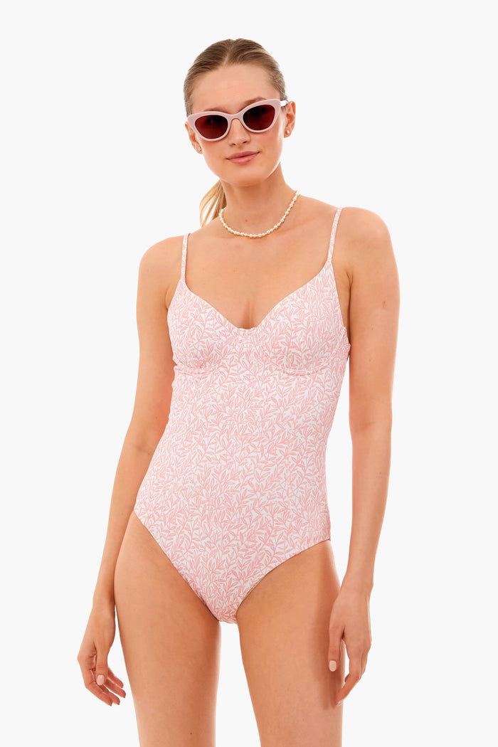 A woman wearing an onia light pink patterned Chelsea one piece swimsuit with four-way stretch fabric, sunglasses, and a pearl necklace stands confidently.