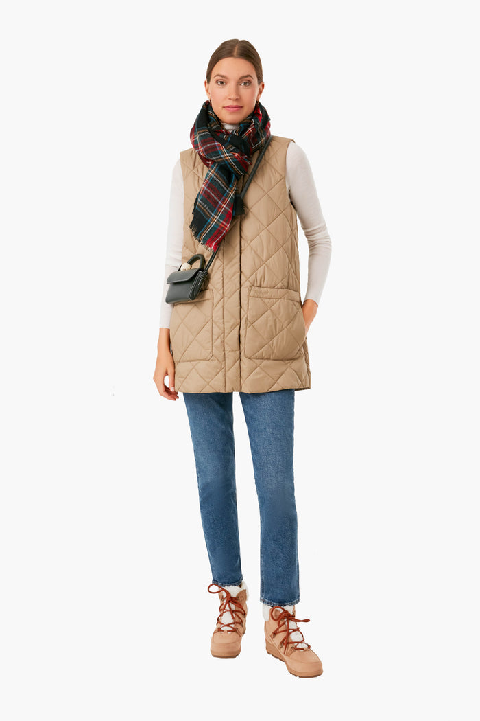 A woman stylishly models Barbour’s beige diamond-quilted vest with a white long-sleeve shirt, plaid scarf, blue jeans, tan boots, and a small black crossbody bag for a chic lightweight outerwear look.