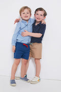 Two boys smile with arms around each other. One wears a blue Oso & Me shirt and adjustable waist kids shorts; the other is in a navy polo and tan shorts. Both complete the look with casual shoes.