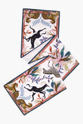 A rectangular silk scarf by Ardmore Design features illustrations of leopards, zebras, gazelles, and tropical plants with a dark border, inspired by Ardmore’s modern collectibles and vibrant South African art.