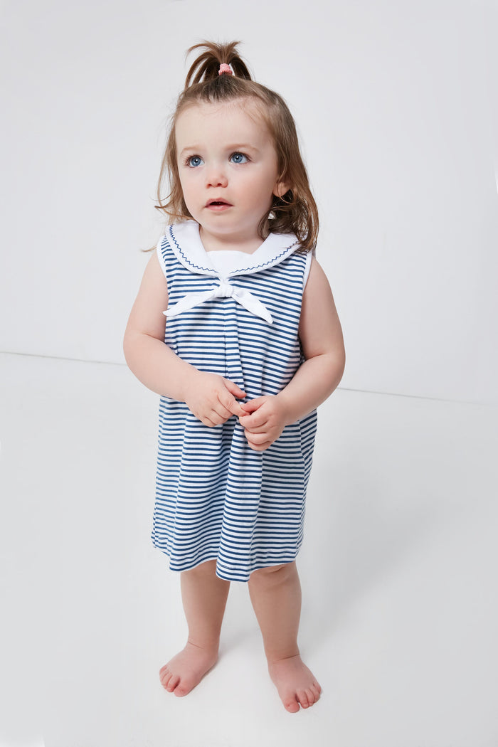 A young child with blue eyes and brown hair wears a sleeveless, blue-and-white striped Pima cotton baby dress by kissy kissy, standing barefoot and highlighting adorable nautical baby clothing.